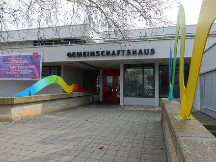Eingang des Gemeinschaftshaus Langwasser, Betongebäude mit roter Metalltür, zum Eingang hin leiten auf einer Brüstung montierte Regenbogen-Schlangen aus lackiertem Blech