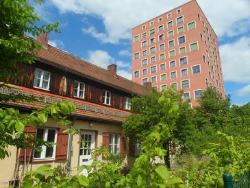 Wohnsituation im Regensburger Viertel im späten Frühjahr bei heiterem Wetter, im Vordergrund Bestandsgebäude aus der NS-Zeit, zweigeschossig mit junger Hainbuchen-Hecke, Fensterläden und kleinen Bäumen; im Hintergrund ragt monolitisch einer der neu erbauten Wohnblocks auf, über zehn Stockwerke, lachsfarben mit weiß umrandeten Fenstern und kupfergrünen Jalousien, ohne Balkone.