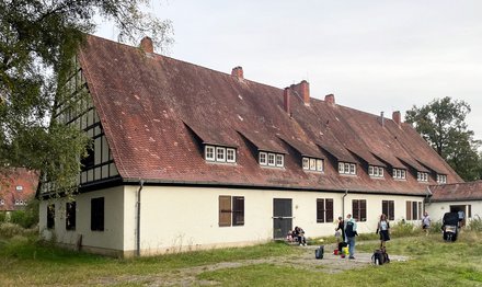 Ansicht des Gebäudes in der Regensburger Straße 406 von seitlich vorne. es ist 2-geschossig und eher länglich mit einem spitzen Dach, Baustil sichtbar nationalsozialistisch. auf der lichten Fläche vor der an der langen Seite gelegenen Eingangstür gehen mehrere Junge Leute, auch ein Fahrradanhänger und Kinder sind dabei. die Eingangstür und die Fensterläden im Erdgeschoß sind geschlossen, aber die Tür im Seitengebäude ist offen, in ihr steht ein junger Mann mit weißem T-Shirt.