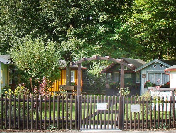 Hinter einem Holzzaun mit hölternem Toreingang eine Gruppe von sechs unterschiedlichen, liebevoll gepflegter Gartenhäuschen, das Laubenmuseum Nürnberg.