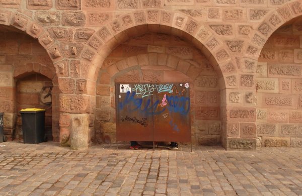 Hinter einer Abschirmung mit Rostoberfläche und Graffitti ist die öffentliche Toilette an/in der Stadtmauer verborgen. Die Wand reicht nicht komplett zum Boden, die letzten ca. 15cm sind offen, dadurch kann man in den beiden Toilettenbereichen  je ein Paar Füße in Turnschuhen erkennen. 