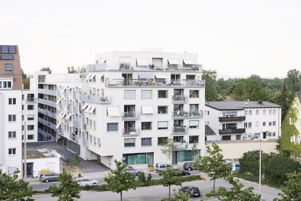 Blick auf die Spiegelfabrik, Nordseite. 6-stöckiges weißes Wohngebäude, unten links der Eingang ist zurückgesetzt, Obere Stockwerke: im Raster angeordnete Fenster und Balkone. An den Fenstern sind teilweise Jalousien heruntergelassen, auf den Balkonen viele viele Topfpflanzen. 