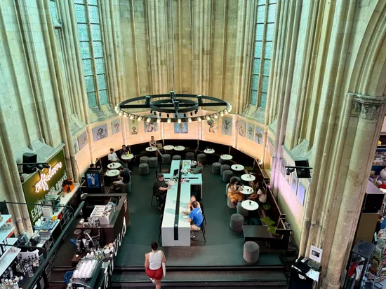 von oben blickt man in den umgebauten Altarraum einer Kirche. Links ist eine moderne Bar mit Neonschriftzug und Essensausgabe; in der Mitte an einem massiveh Tisch in Form eines liegenden Kreuzes sitzen einzelne Menschen mit Tellern, aussen um diesen großen Tisch stehen in einer Reihe viele kleine runde weiße Cafétische und puschelige graue Hocker, auch hier sitzen Menschen mit Tellern. In der Mitte des Raumes hängt ein massiver Leuchter, die Kirchenfenster lassen im oberen Bereich grünliches Licht ein, während im unteren Bereich warmes Licht dominiert. An der Wand sind gemalte Portraits aufgehängt.