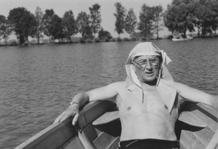 Historisches Foto: In einem Holz-Ruderboot auf einem See lehnt entspannt Alfred Hensel, ein älterer, weißer Mann mit Brille. Er trägt eine Badehose und hat sich ein dünnes Baumwolltuch als Sonnenschutz auf den Kopf drapiert. Im Hintergrund ist eine Baumreihe zu sehen, vielleicht Südseite des Dutzendteichs. 