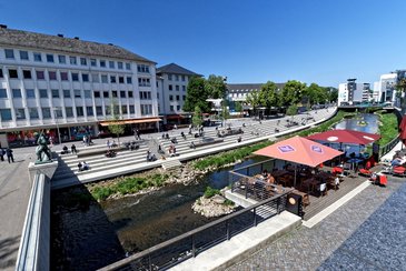 Weitwinkliges Foto: Blick aufs Siegufer, im Vordergrund eine Art Balkon über dem Fluß mit Sonnenschirmen und Gastronomie, links führt eine Brücke über den Fluss, auf der gegenüberliegenden Seite gutbesuchte Sitzstufen, vom Straßenniveau bis hinunter zum Fluss, ca. 200m am Fluss entlang bis zur nächsten Brücke.