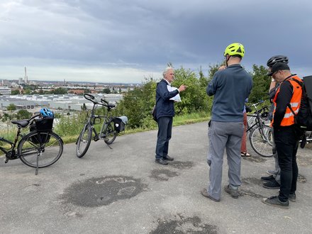 Werner Geim mit gedruckter Broschüre steht auf einem sichtbar in die Jahre gekommenen reparierten geteerten Platzoben auf dem Schweinauer Buck vor einer Gruppe von 5 Leuten, zum Teil mit Fahrradhelmen, die ihm interessiert zuhören. Hinter ihm sind Fahrräder der Gruppe abgestellt, im Hintergrund hat man Aussicht über Schweinauer Industrieanlagen, Müllverbrennungsanlage, Südstadt,Stadt und Burg.