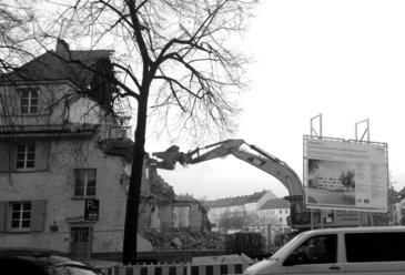 Die Schaufel eines Abrissbaggers, geformt wie ein Kopf mit einem Maul, beisst an einem gar nicht so kaputt aussehenden 3-stöckigen Eckhaus aus den 1920ern herum, vom früheren Wohnblock sind schon drei Viertel weg. 