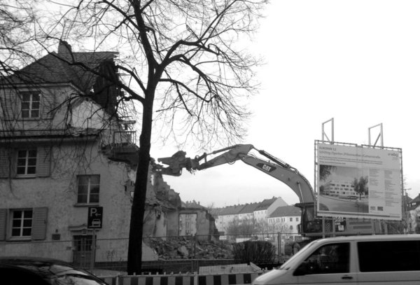 Die Schaufel eines Abrissbaggers, geformt wie ein Kopf mit einem Maul, beisst an einem gar nicht so kaputt aussehenden 3-stöckigen Eckhaus aus den 1920ern herum, vom früheren Wohnblock sind schon drei Viertel weg. 