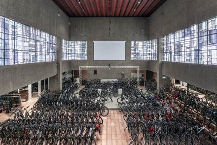Foto in eine große Halle, ein moderner großer Raum, oben durch ein Band von Glasfenstern in blau-grau Tönen diffus beleuchtet, unten stehen ordentlichst aufgereiht und durch Mittelgang geteilt ca 200 neue Fahrräder