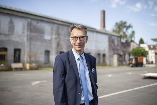 Daniel Furhop, mit Anzug und Brille, im Hintergrund unscharf ein geteerter Platz, dahinter ein in die Jahre gekommener Industriebau, an dessen Hinterseite ein Gabelstapler zu sehen ist, weiter vorne aber aus Paletten gebaute Beete und eine Sitzbank.