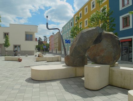 Zentraler Platz im Regensburger Viertel, mehrere Steinskulpturen, unten weiße Kalkstein-Riegel, auf denen man auch sitzen kann, auf denen einige dunkle Findlinge miteinander kombiniert sind (Höhe 40cm–2m). der Platz gepflastert. rechts im Hintergrund 4-geschossige, farbige Gebäude der Blockränder mit weiß umrandeten Fenstern, links ein 2-geschossiges weißes Gebäude. 