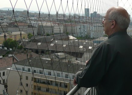Von einem Aussichtspunkt geht der Blick über die Dächer Wiens. Rechts im Bild Manfred Jupitz, ein grauhariger Mann mit Brille, blickt interessiert und angeregt auf die unter ihm liegende Stadt.