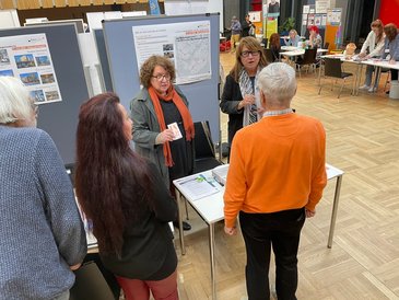 Brigitte Sesselmann und und Ulla Krämer hinterm Stand unterhalten sich angeregt drei Personen, die Atmosphäre ist freundlich und zugewandt.