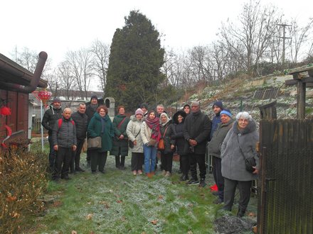 17 Personen, in dicken Jacken haben sich zwischen dem Bahndamm und der Wand einer Gartenlaube auf die vereiste Wiese zum Gruppenfoto aufgestellt. Sie blicken ernst in die Kamera. Die Gruppe ist sehr gemischt: einige sind alt, aber es ist auch eine Familie mit Jugendlichen dabei. In der Mitte der Gruppe zwei Frauen von BauLust und NFSK.