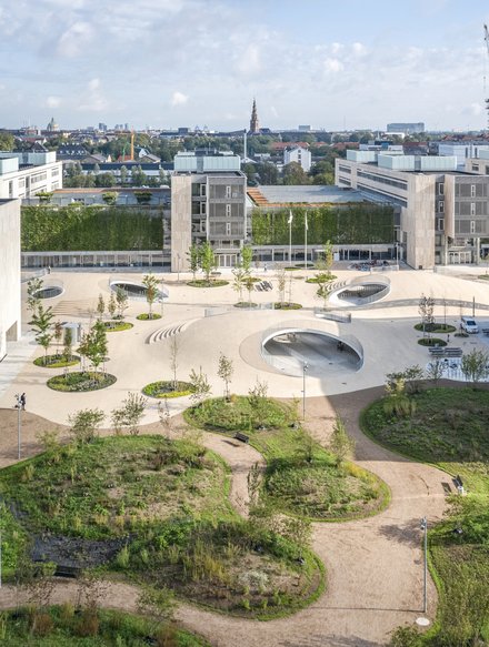 Luftbild des Karen-Blixens-Platz im vorderen Teil ein Park mit durchlässigen Wegen und runden Pflanzeninseln, im hinteren Teil großteils versiegelt, aber durchsetzt von runden Pflanzeninseln, dreimal erheben sich begehbare Hügel/Kuppeln mit runden Eingangs-Durchbrüchen, innen Fahrradständer, aussen Brücken und Sitzgelegenheiten