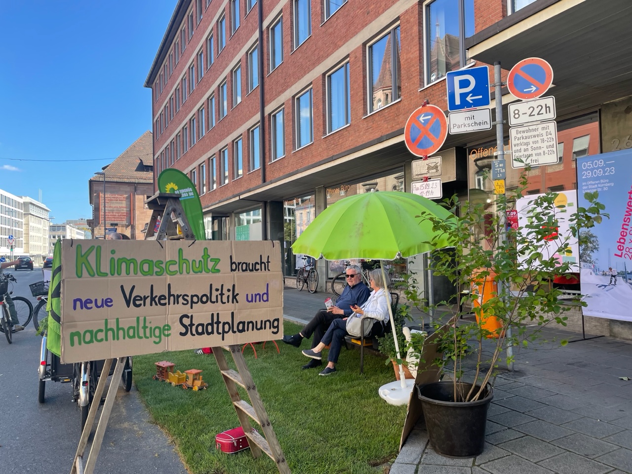 Mit Rollrasen zur Grünfläche umgestaltete Parkplätze vor dem Offenen Büro, als Begrenzung aufgestellt eine Leiter mit Demonstrationsplakat aus Karton – Klimaschutz braucht neue Verkehrspolitik und nachhaltige Stadtplanung – auf der rasenfläche ein Sonnenschirm und eine Bank, auf der Bank Sepp und Agnes Reindl