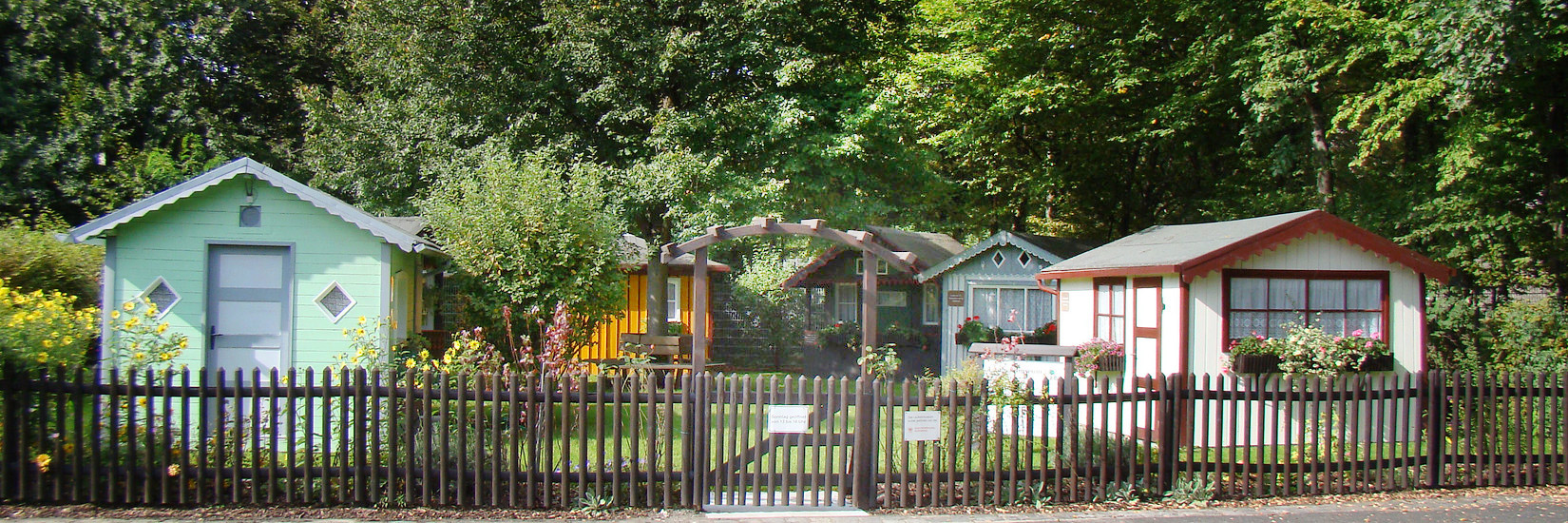 Hinter einem Holzzaun mit hölternem Toreingang eine Gruppe von sechs unterschiedlichen, liebevoll gepflegter Gartenhäuschen, das Laubenmuseum Nürnberg.