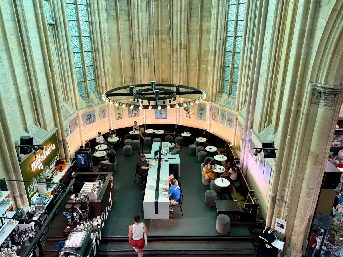 von oben blickt man in den umgebauten Altarraum einer Kirche. Links ist eine moderne Bar mit Neonschriftzug und Essensausgabe; in der Mitte an einem massiveh Tisch in Form eines liegenden Kreuzes sitzen einzelne Menschen mit Tellern, aussen um diesen großen Tisch stehen in einer Reihe viele kleine runde weiße Cafétische und puschelige graue Hocker, auch hier sitzen Menschen mit Tellern. In der Mitte des Raumes hängt ein massiver Leuchter, die Kirchenfenster lassen im oberen Bereich grünliches Licht ein, während im unteren Bereich warmes Licht dominiert. An der Wand sind gemalte Portraits aufgehängt.